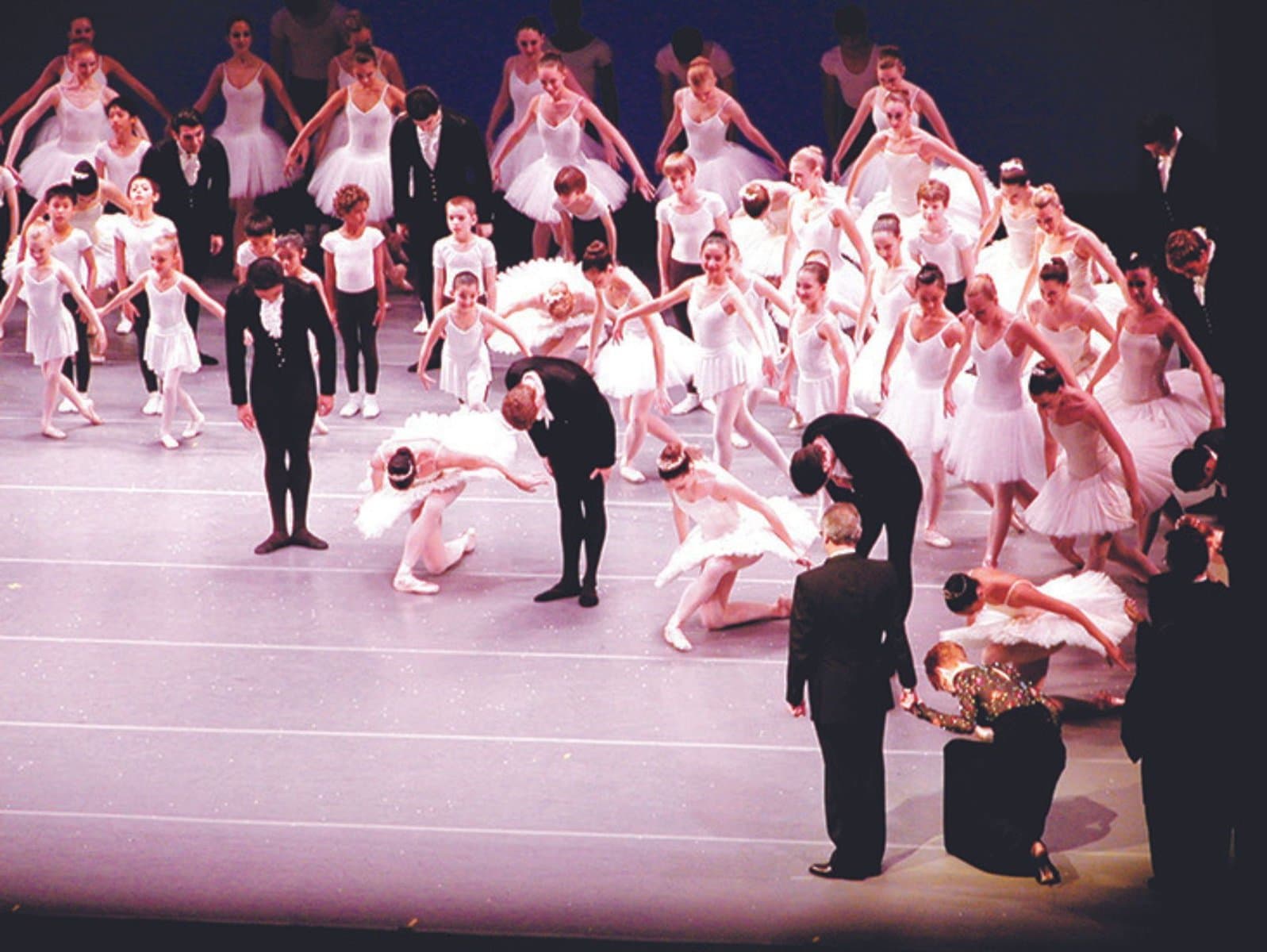 On stage, a large group of ballet dancers and students, dressed in black and white, bow towards Kent Stowell and Francia Russell. Francia is in a deep curtsy.