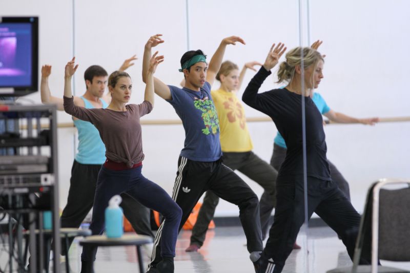 A group of dancers rehearse in a spacious studio with mirrored walls. They are dressed in comfortable athletic wear. The dancers appear focused, with their arms raised gracefully and legs positioned in a dynamic stance. A woman in black guides the group.