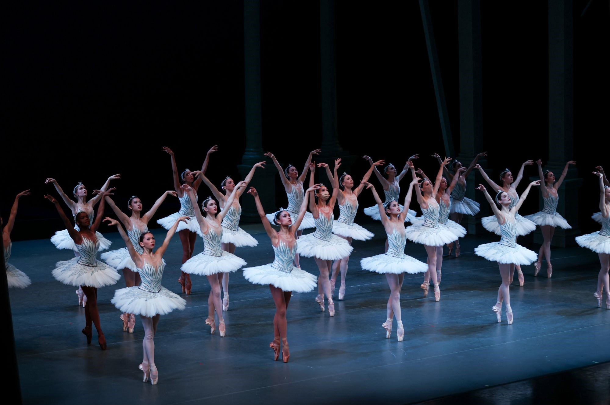 A large group of dancers wearing white tutus stand in rows on a darkly lit stage. They are looking up towards the sky with arms reaching up.