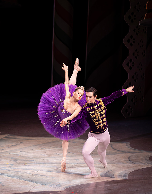 A dancer in a glittery tutu reaches across the stage and lifts her leg behind her while another dancer in a velvet purple coat supports her.