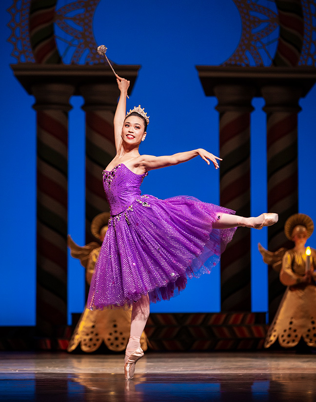 A dancer in a purple sparkly tutu stands in arabesque.