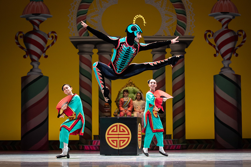 A dancer in a black unitard with green accents leaps on stage with arms outstretched.
