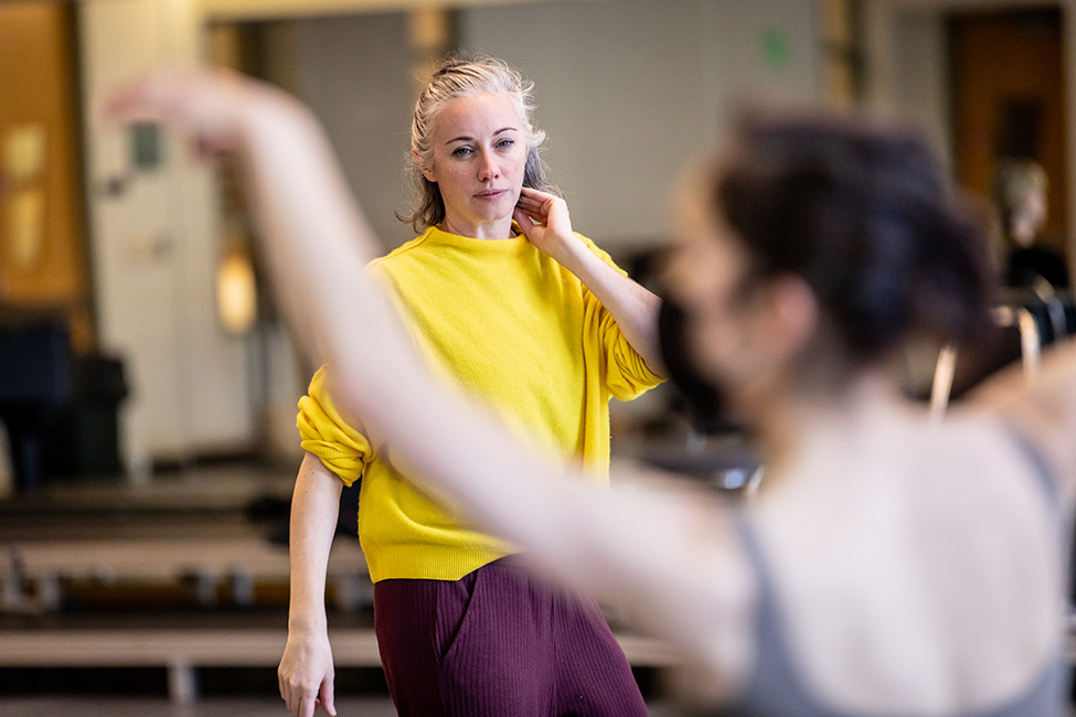 Dani Rowe, wearing a yellow sweater, carefully observes a dancer in rehearsal.