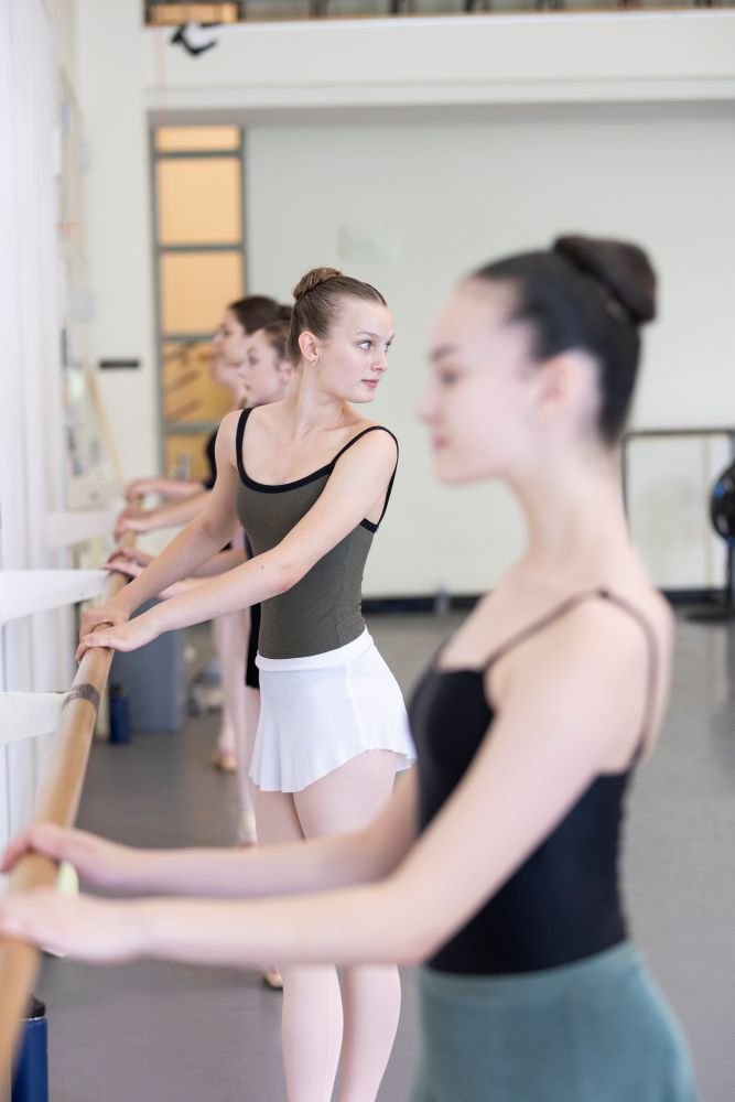 Three dancers stand facing the barre in a studio with white walls. One looks over her shoulder, observing the teacher who is out of sight.