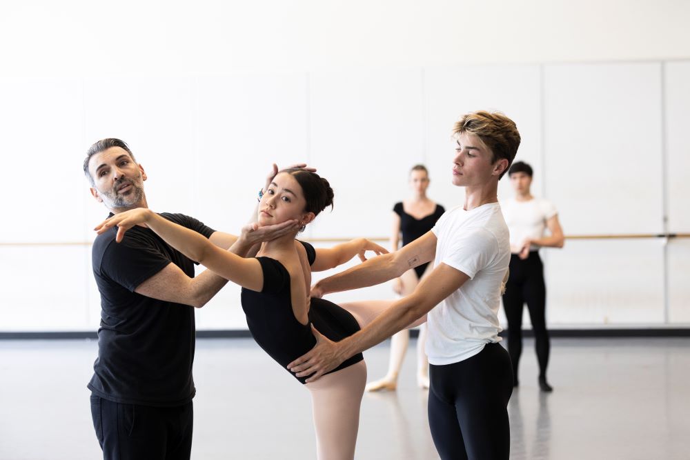 A dance teacher assists a student who is wearing a black leotard and is standing in arabesque.