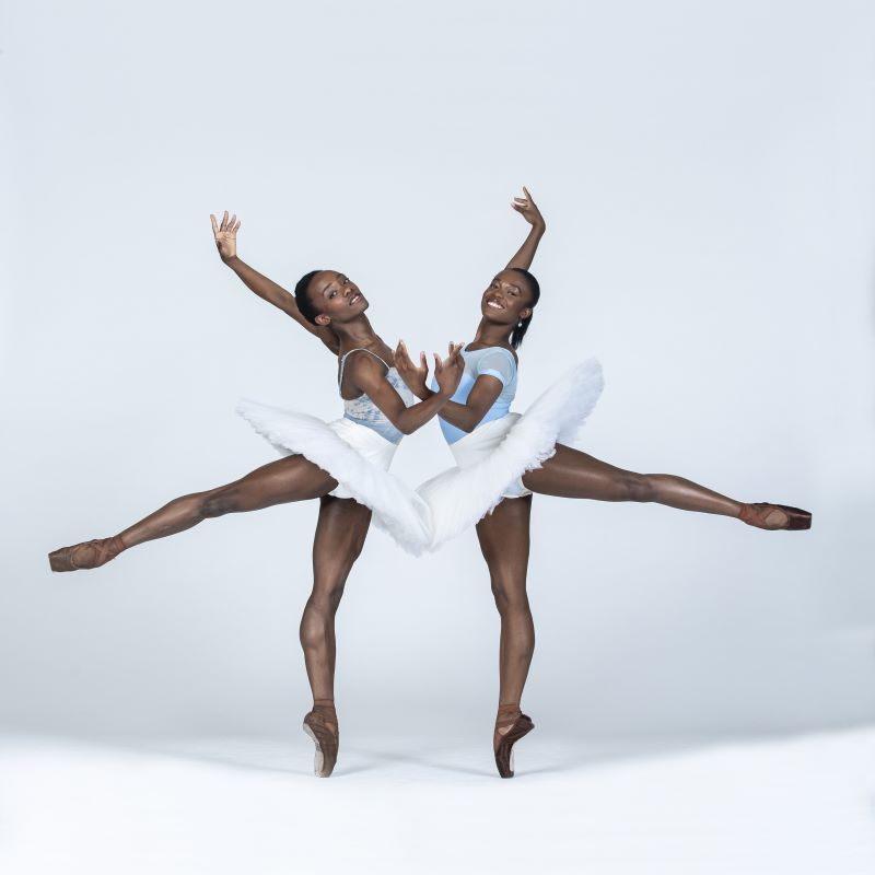 Ashton Edwards and Destiney Wimpey, both in blue leotards and white tutus, stand in arabesque mirroring one another.