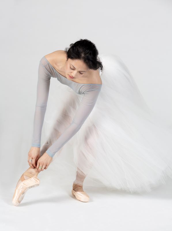 Leah Terada stands in a long white skirt, and leans over to re-tie her pointe shoe.