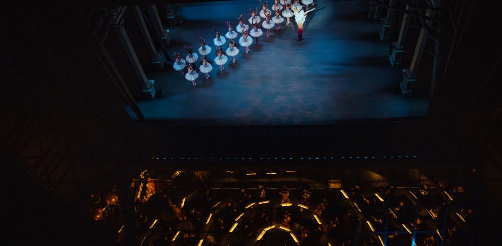 A large group of dancers in white tutus stand on stage with the glowing orchestra pit beneath them.