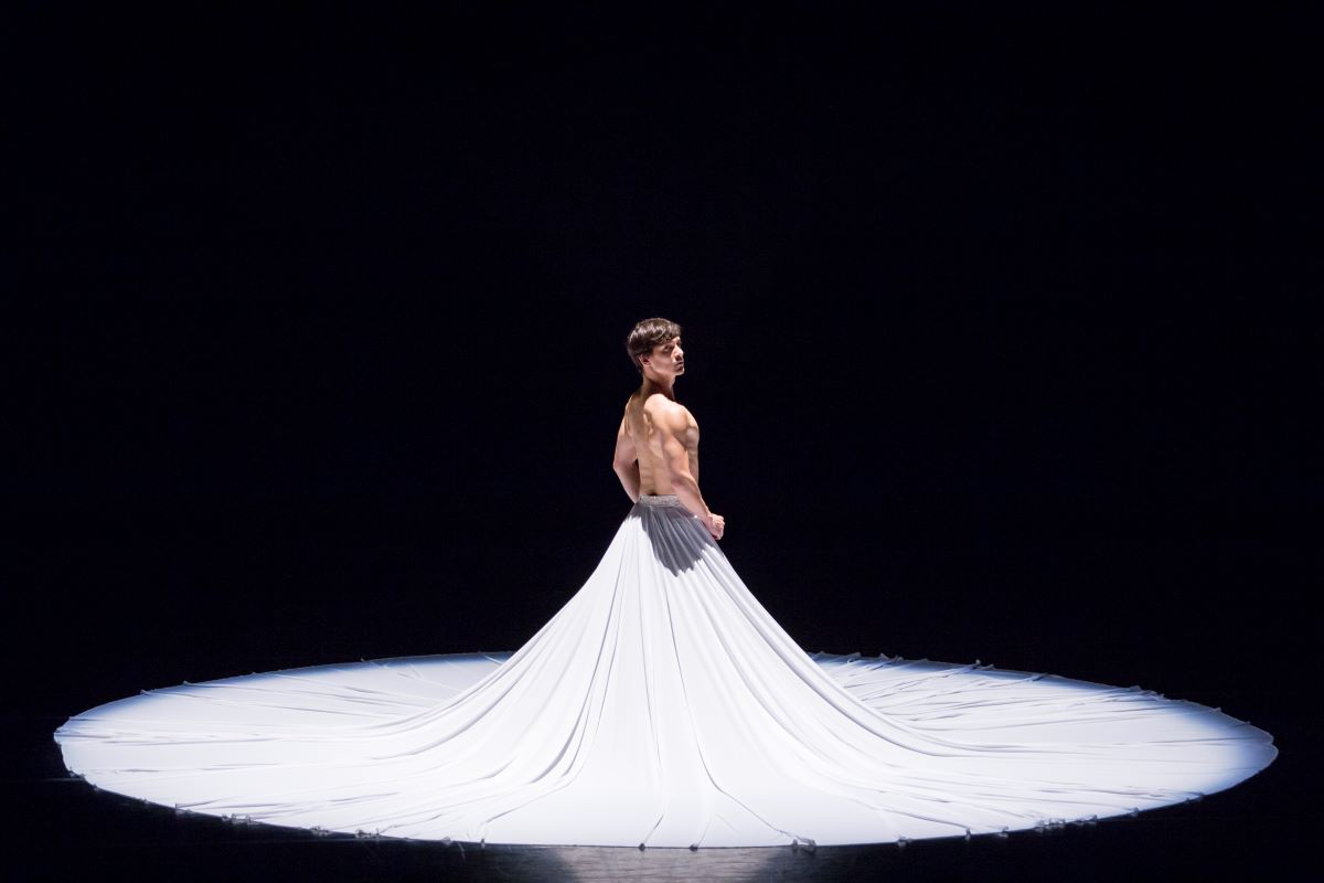 A dancer stands looking over their shoulder on a black stage in a long, dramatic white skirt.