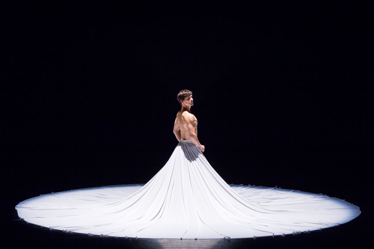 TheCalling2015_Moore_LT_01-websize A dancer stands looking over their shoulder on a black stage in a long, dramatic white skirt.