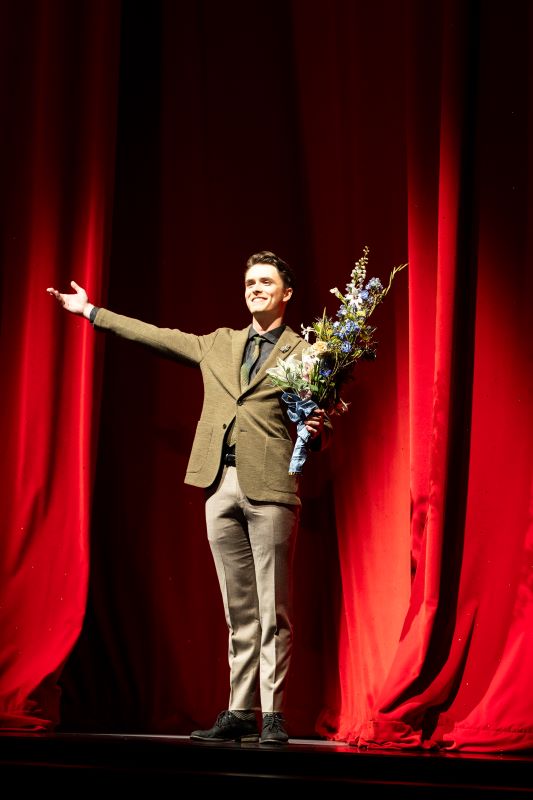 Rep1.Open 097 A smiling Luther DeMyer stands in front of a red, sparkly curtain. His arms are reaching out, and he is holding a bouquet of flowers.