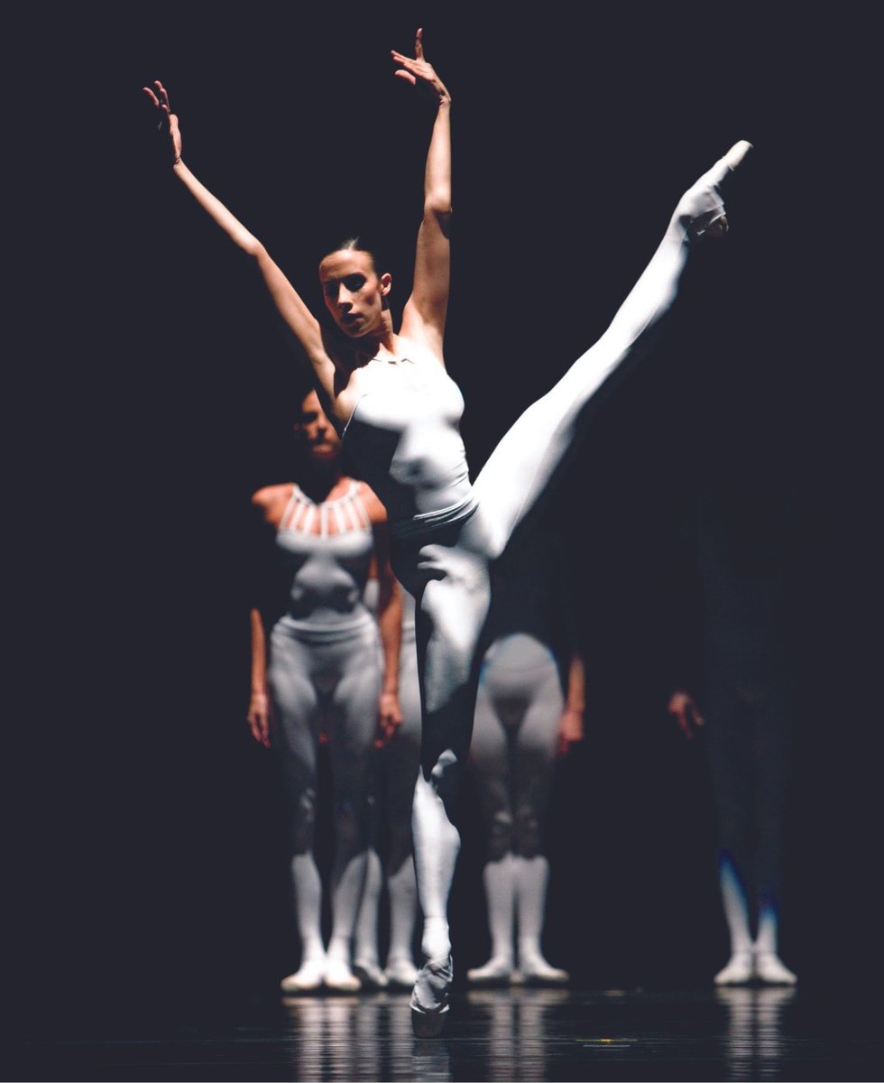 Mid-performance, a dancer reaches both arms overhead as she extends her leg up and to the side. She is wearing a white unitard which is striking against the black background.